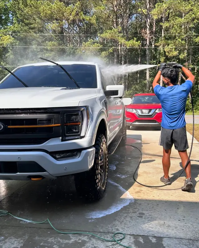 A BMC crew member pressure-rinsing a white Ford F-150 on a concrete driveway